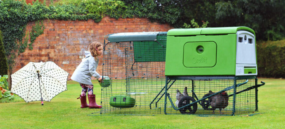 Ein Mädchen füttert ihre Hennen in einem grünem Eglu Cube Hühnerstall mit Auslauf