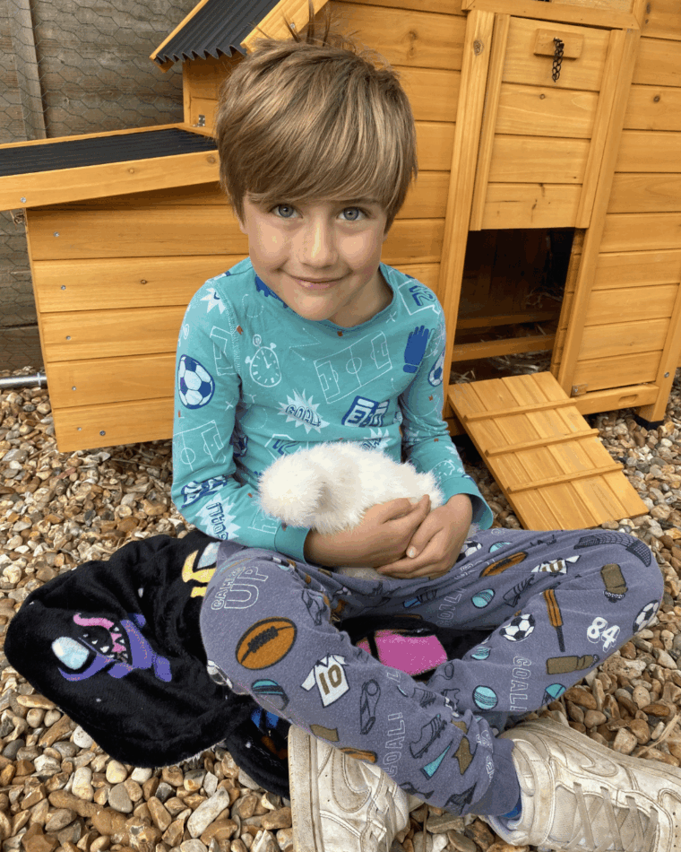 Ein kleiner Junge sitzt mit einem Huhn auf dem Schoß vor dem Hühnerstall